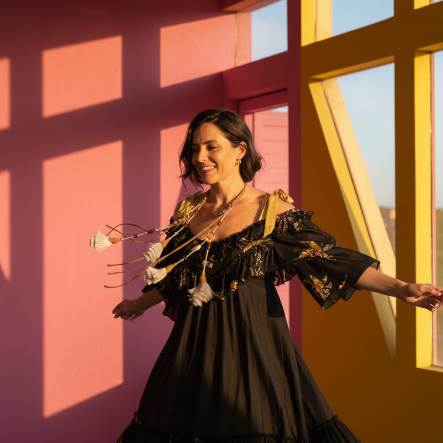 A black cold-shoulder blouse with gold floral prints and cream ribbon ties, displayed on a white mannequin torso. The blouse is styled with a long bohemian-style beaded necklace featuring white tassels.