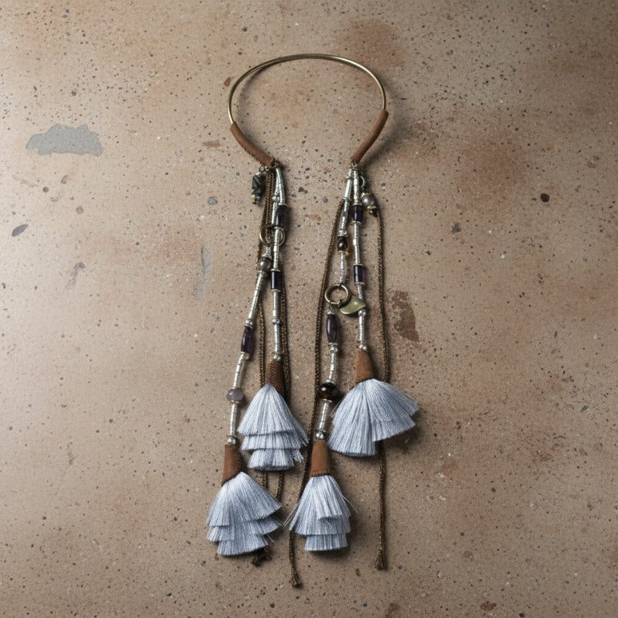 A pair of long bohemian-style dangle earrings with layered silver-gray tassels, dark beads, and metallic brown details, displayed on a distressed beige fabric background.