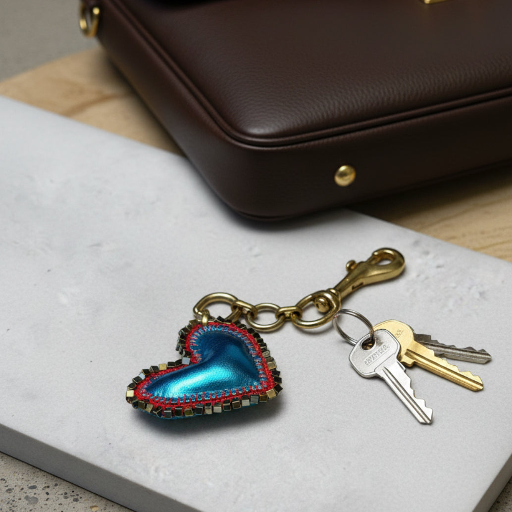 A heart-shaped keychain charm with a metallic blue center, red and silver beaded border, and an antique gold clasp, displayed on a white marble tile.
