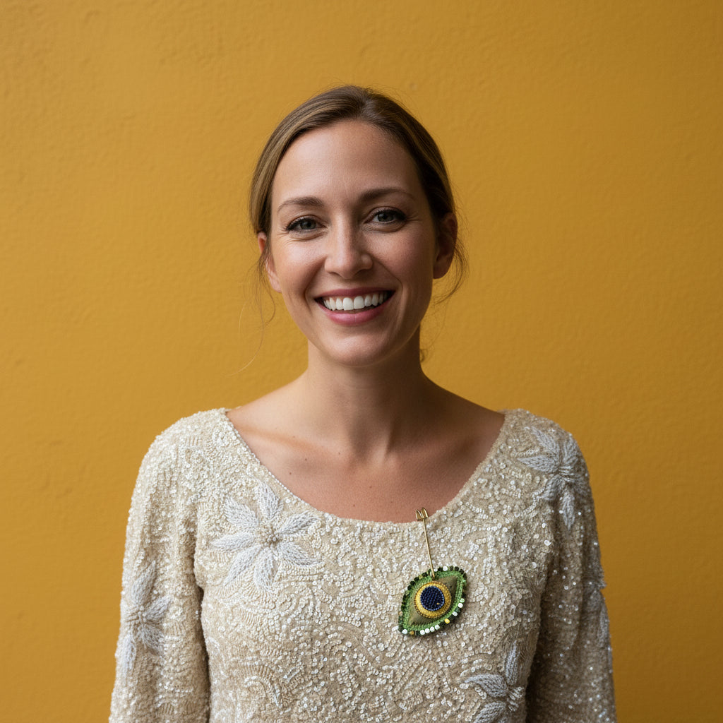 A close-up of a cream-colored beaded blouse with floral embroidery, displayed on a white fabric mannequin torso. A decorative green and gold beaded pin is pinned to the chest area of the blouse.