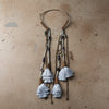 A pair of long bohemian-style dangle earrings with layered silver-gray tassels, dark beads, and metallic brown details, displayed on a distressed beige fabric background.