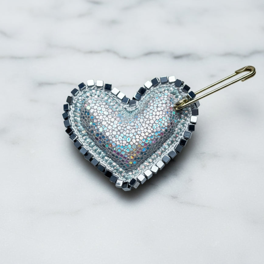 Heart-shaped brooch with metallic and iridescent design on a textured fabric background