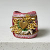 A handmade cuff bracelet featuring a large gold heart charm, gold metal beads, silver and pink sequins, green velvet fabric, and white ribbon on a neutral light gray background.