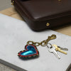 A heart-shaped keychain charm with a metallic blue center, red and silver beaded border, and an antique gold clasp, displayed on a white marble tile.