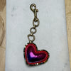 A heart-shaped keychain charm with a metallic purple and pink center, red stitched border, and a bronze-toned clasp, displayed on a white rectangular surface.