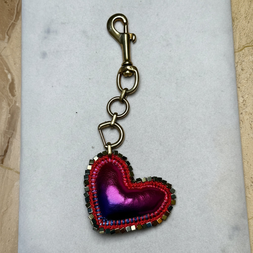 A heart-shaped keychain charm with a metallic purple and pink center, red stitched border, and a bronze-toned clasp, displayed on a white rectangular surface.