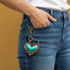 A person wearing mustard yellow corduroy shorts and a gray top, shown from the waist down in a home setting. A heart-shaped pendant necklace with a blue center and gold border hangs from the shorts pocket.