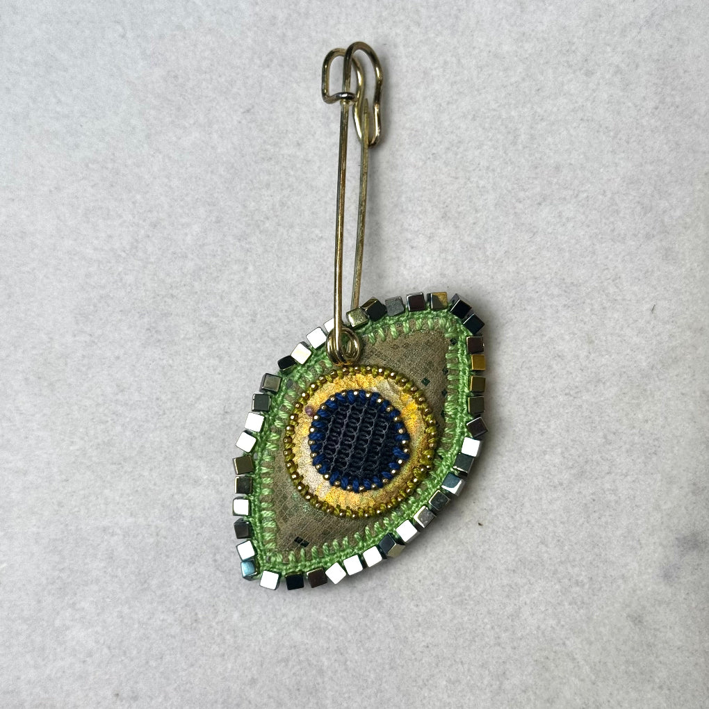 A handmade beaded safety pin accessory featuring a green and gold leaf-shaped pendant with a blue and yellow center, displayed on a light gray textured background.